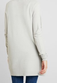 Světle šedý pletený svetr s ribovanou strukturou, dlouhými rukávy a mírně prodlouženým zadním lemem. Zvýrazněn nařasenými manžetami.