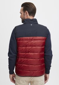 Gilet rembourré rouge et marine avec un design matelassé, col haut et fermeture éclair. Texture lisse avec un tissu contrastant sur les épaules et le dos.