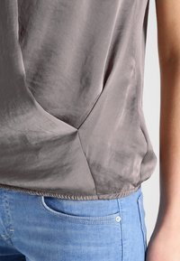 Close-up van een persoon die een losse, grijze satijnen top draagt, ingestopt in lichtblauwe denim jeans met zichtbare voorzak en stikseldetails.