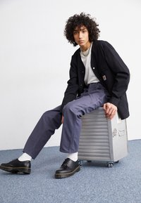 Black jacket over a white t-shirt, gray trousers, black shoes with a thick sole, sitting on a silver rolling suitcase in a minimal setting.