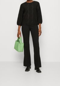 Blusa nera texturizzata con motivi ondulati, abbinata a pantaloni neri aderenti e una borsa verde opaca. Modella che indossa stivali neri chunky.