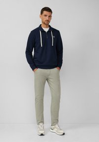 Sudadera de forro polar azul marino con cremallera y cordones blancos, combinada con pantalones ajustados verde claro y zapatillas beige con un patrón texturizado.