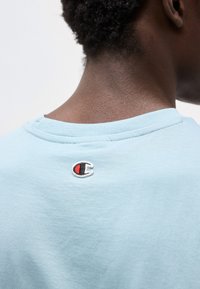 Πίσω όψη ενός ατόμου που φοράει ένα ανοιχτόγαλαν μπλουζάκι με το μικρό λογότυπο της Champion κεντημένο στο πάνω μέρος του πίσω μέρους του λαιμού.