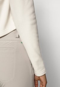 Lättvikts fleece-overall med synliga sömmar, kombinerad med beige slim-fit byxor med bakfickor och en liten grön detaljtag.