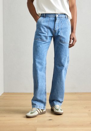 Mann trägt hellblaue Jeans mit geradem Bein und aufgesetzten Fronttaschen, weißes T-Shirt und weiße Sneaker mit braunen und grünen Streifen.
