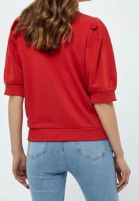 Žena v červeném topu s nafouknutými rukávy a světle modrých džínách, viděná zezadu s vlnitými hnědými vlasy padajícími přes ramena.