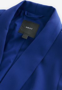 Blue blazer with a smooth, textured fabric, featuring a sharp collar and inner label reading "NEXT." Simple and tailored design.