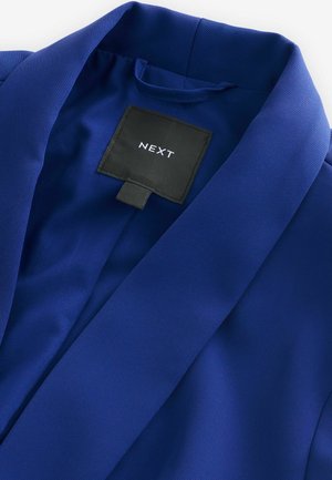 Modrý blazer s hladkým, texturovaným materiálem, s ostrým límcem a vnitřním štítkem s nápisem "NEXT." Jednoduchý a přizpůsobený design.