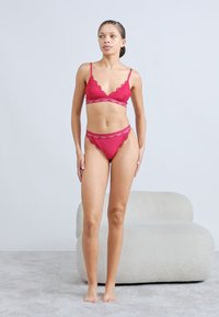 Rotes Spitzen-Bralette mit gewellten Kanten und passendem Tanga. Verstellbare Träger und Logo-Band-Detail. Glatte Textur vor einfarbigem Hintergrund.