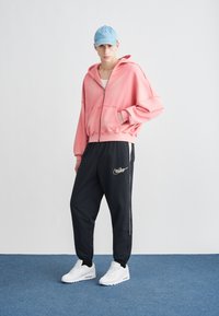 Ροζ φούτερ με φερμουάρ και τσέπη καγκουρό, συνδυασμένο με μαύρη φόρμα με χρυσό λογότυπο Nike και κάθετες ρίγες στο πλάι, φορεμένο με λευκά αθλητικά παπούτσια.