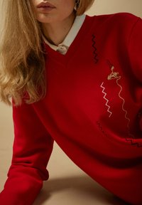 Sieviete ar gariem blondiem matiem, valkājot sarkanu džemperi ar zigzaga raksta dūrienu virs baltas apkaklītes krekla neitrālā fonā.