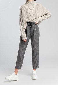 Beige-värinen palmikkoneulepooloneule, yhdistetty harmaisiin ruudullisiin kapeamalahkeisiin housuihin, joissa on joustava vyötärönauha, ja valkoisiin tennareihin.