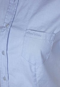 Camicetta azzurra chiara in tessuto morbido, con taschino che presenta la scritta "Marc O'Polo" in rilievo e dettagli dei bottoni ben visibili.
