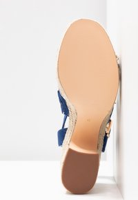 Semelle d'une sandale compensée bleue avec une semelle intermédiaire en jute et un talon en caoutchouc texturé, taille 9, présentée sur fond blanc et dans un coin de mur.