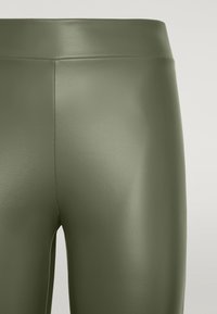 Olivgröna leggings i konstläder med en slät textur, hög midja och synliga sömdetaljer. N ärbild som visar materialkvaliteten.