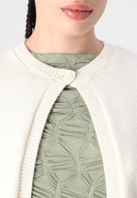 Cardigã de malha creme com bordas caneladas e uma gola redonda, fechado com um botão ao centro, sobre uma blusa texturizada verde-oliva.