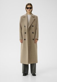 Abrigo de mezclilla de lana beige de doble botonadura con grandes botones negros, solapas amplias y bolsillos laterales, usado sobre pantalones grises y zapatos negros.