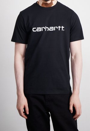 Mand iført en sort Carhartt t-shirt med hvidt logo, kombineret med sorte bukser, står foran en ensfarvet lys baggrund.