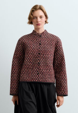Femme aux cheveux blonds courts porte une veste boutonnée à manches longues avec un motif rouge et noir et un pantalon large noir.