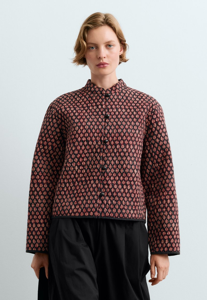 Femme aux cheveux blonds courts porte une veste boutonnée à manches longues avec un motif rouge et noir et un pantalon large noir.