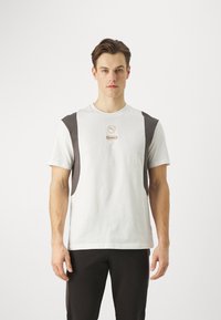 Witte katoenen t-shirt met donkergrijze schouderaccenten, met een klein lichtbruin logo gecentreerd op de borst. Eenvoudige ronde hals.