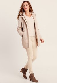Manteau matelassé beige avec une capuche bordée de fourrure, poches latérales, associé à un pull léger et un jean beige, complété par des bottines brunes.