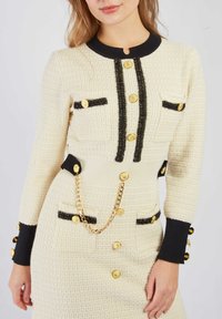 Biała teksturowana sukienka z czarnymi akcentami, złotymi guzikami i detalem w postaci łańcucha. Posiada zaokrąglony dekolt i dopasowany krój z kieszeniami.