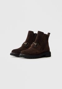 Une paire de bottines en daim marron foncé avec des semelles noires épaisses et un détail en métal sur le devant, dotées de fermetures éclair sur les côtés et de languettes de tirage.
