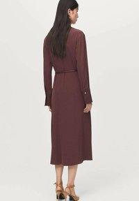 Abito midi bordeaux con maniche lunghe, dettaglio con cintura legata e polsini con bottoni. Tessuto morbido con una consistenza delicata. Indossato con sandali con tacco color tan.