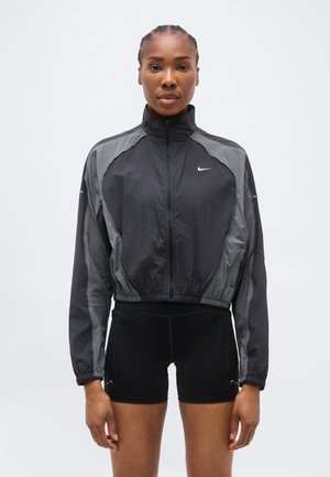 Naine seisab rinna ees, seljas musta ja halli Nike lukuga jope ning mustad spordisortsid, taustaks lihtne valge sein.