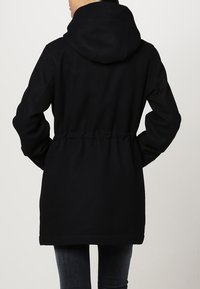 Zwarte capuchonjas met een getailleerde taille, gemaakt van een zachte stof. Voorzien van lange mouwen en een rechte zoom ter hoogte van de heupen.