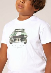 T-shirt blanc avec un motif de voiture vintage verte avec des détails noirs, des rayures blanches verticales et le texte "Dee Luxe Original."