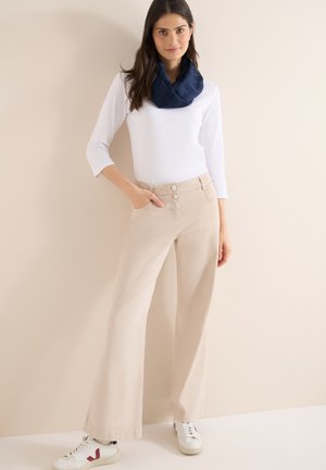 Femme debout portant un haut blanc à manches trois-quarts, un pantalon beige à jambes larges, des baskets blanches et une écharpe bleu marine sur un fond beige uni.