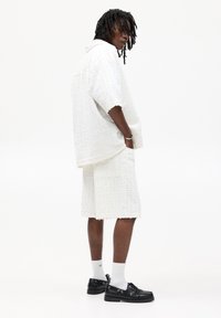 Set di camicia corta a maniche corte bianca con texture e pantaloni a shorts. Presenta una vestibilità rilassata, bordi grezzi ed è abbinato a scarpe nere e calzini bianchi.