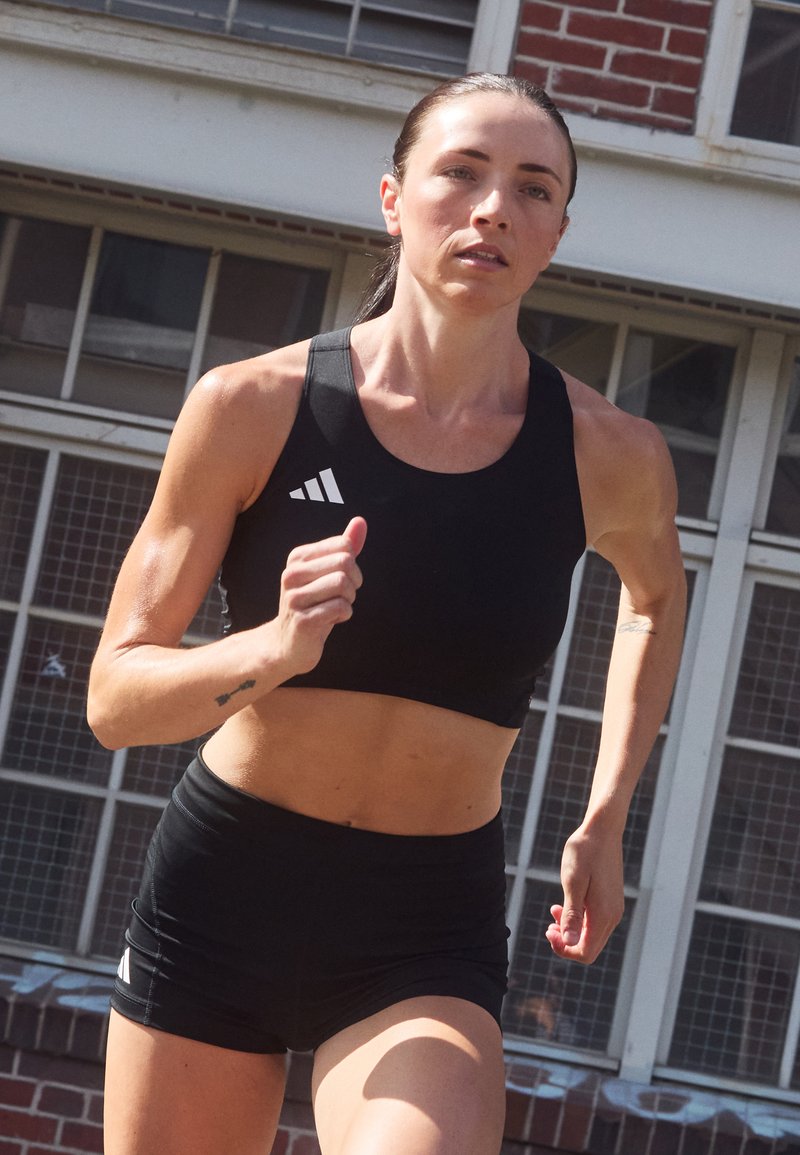 Svart atletisk crop top och shorts i fukttransporterande material. Har vita logotypdetaljer. Modellen springer i en utomhusmiljö.