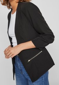 Blazer noir à coupe ajustée, manches longues, avec une poche zippée sur le bas devant. Porté sur un haut blanc et un jean bleu.