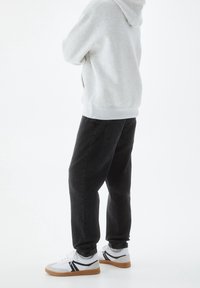 Osoba ubrana w jasnoszarą bluzę z kapturem, luźne czarne dżinsy i białe trampki z gumową podeszwą, stojąca na białym tle.