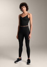Fekete sportos tank top vékony pántokkal, magas derékú fekete leggingszel párosítva. Az anyag rugalmasnak és simának tűnik. Fekete sportcipők.