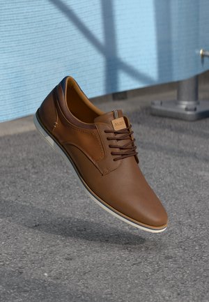 Brown leather shoe with a textured finish, lace-up design, white sole, and subtle stitching details. Floating above a gray surface.