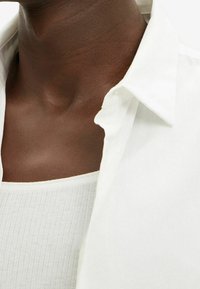 Camisa blanca de cuello con algunos botones desabrochados sobre una camiseta de tirantes de un gris claro con textura acanalada. Primer plano del escote y el hombro, con una textura de piel suave visible.