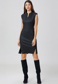Robe noire sans manches avec un col montant, des fentes sur les côtés et une texture lisse ; assortie à des bottes en daim vert foncé montantes jusqu'aux genoux.