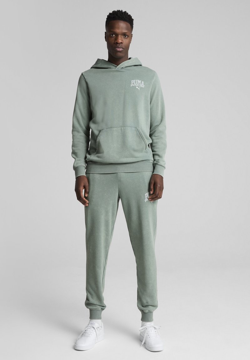 Felpa con cappuccio grigio-verde in pile con tasca a marsupio, abbinata a pantaloni della tuta con polsini coordinati. Entrambi presentano un piccolo logo bianco sul petto e sulla coscia.