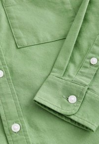 Chemise en tissu vert avec une poche, au texture lisse, boutons blancs et un design de poignet replié.