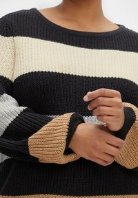Persona che indossa un maglione a maglia con ampie righe orizzontali in nero, beige, grigio e crema, con le mani piegate vicino alla vita.