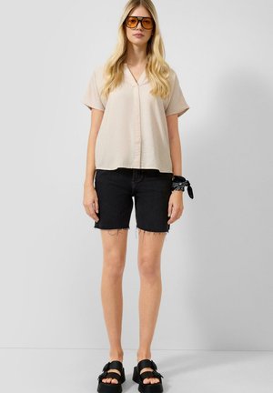 Femme aux longs cheveux blonds portant un chemisier beige à manches courtes, un short en jean noir, des sandales noires, des lunettes de soleil ambrées et un bandana noir au poignet.