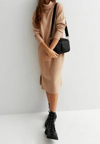 Beiges Strickkleid mit Rollkragen, gerippten Bündchen und einem seitlichen Schlitz, kombiniert mit schwarzen Leder Schnürstiefeln und einer kleinen schwarzen Umhängetasche.