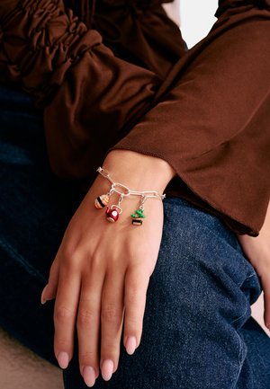 Hand mit braunem Ärmel und blauer Jeans, geschmückt mit einem silbernen Charm-Armband mit Anhängern in Form von Sushi, Teekanne und Kaktus.