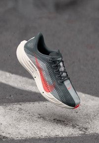 Chaussure de sport grise avec une tige en maille légère, des accents rouges, une semelle arrondie et le logo emblématique de Nike. Conçue pour la performance.