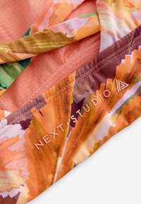 Blomsteret stof med klare orange og pink nuancer, med en struktureret ferskenfarvet baggrund og trykt "NEXT STUDIO" branding.