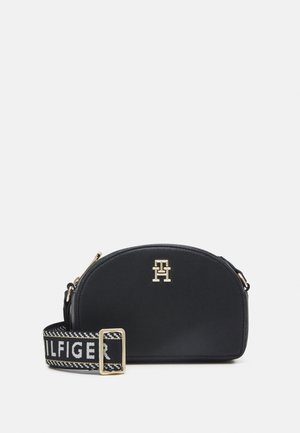 Sac bandoulière en cuir noir de forme arrondie, avec des éléments métalliques dorés et une sangle à motif arborant le logo "TOMMY HILFIGER".